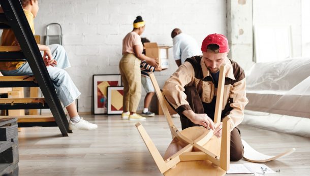 Young furniture assemblage specialist in workwear standing on knees on the floor and assembling new wooden chair against settlers
