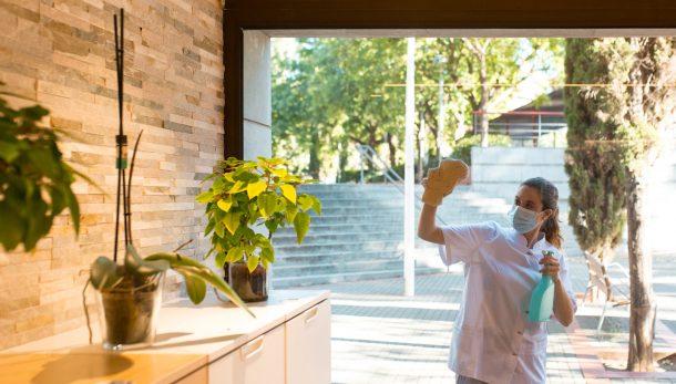 Front view of young woman in uniform and wearing a face mask cleaning windows of spa center from outside.